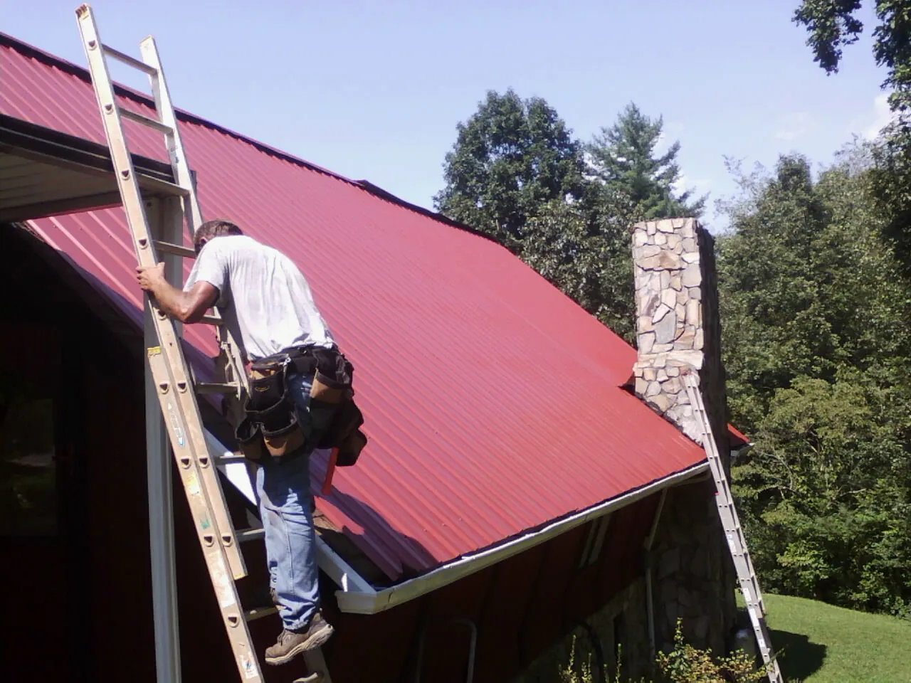 Roofer on a ladder beside a red metal roof and stone chimney — Soffit & Fascia Repair by Roofing Brothers in Hurst